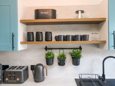 A kitchen countertop with a toaster kettle and hanging plants under wooden shelves with jars mugs and a bread box at Savoy View in Mullion