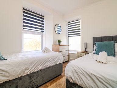 A bedroom with two single beds a chest of drawers a small table with a lamp and windows with striped blinds at Savoy View in Mullion