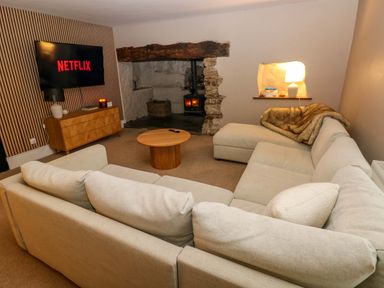 A living room with a beige sectional sofa a round wooden coffee table a wooden cabinet with a lamp and candle a wall-mounted tv showing Netflix and a stone fireplace with a fire at Norchard Farm House in Redberth near St Florence