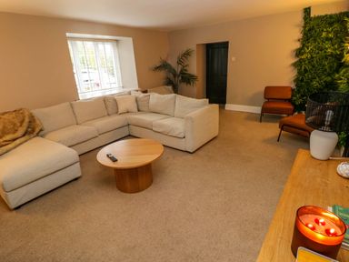 A living room with a beige sectional sofa a round wooden coffee table two orange chairs a large window and a green wall panel at Norchard Farm House in Redberth near St Florence