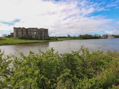 A ruined castle near a body of water with bushes in the foreground and farmland in the background