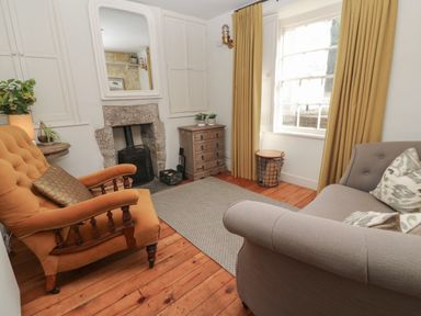 A living room with an orange armchair a gray sofa a stone fireplace wooden floor and a window with yellow curtains at Apple Tree Cottage in Gweek