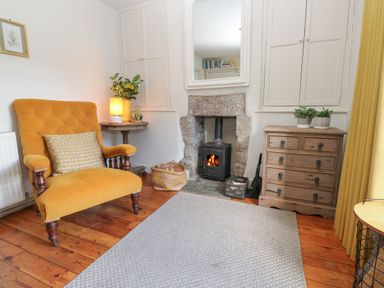A room with a yellow armchair a small round table with a lamp plants a stone fireplace with a wood stove a wooden chest of drawers and a gray rug at Apple Tree Cottage in Gweek