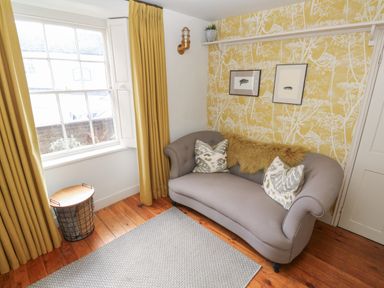 A small living room with a gray sofa two patterned cushions yellow curtains wooden floor and a patterned yellow wall at Apple Tree Cottage in Gweek