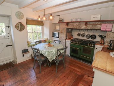 A kitchen with a round table and chairs an oven hanging pots knives and a window seat at Apple Tree Cottage in Gweek