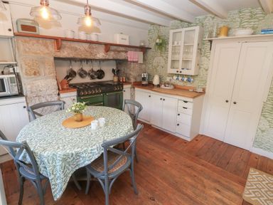 A kitchen with a round table covered with a patterned tablecloth and four chairs next to a stove set in a stone alcove and white cabinets at Apple Tree Cottage in Gweek