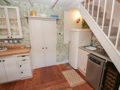 A kitchen area with wooden floor, white cabinets, a refrigerator, dishwasher, sink under stairs at Apple Tree Cottage in Gweek