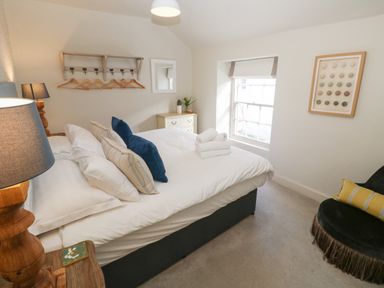 A bedroom with a bed with pillows and folded towels a window nightstand with a lamp a black chair and a chest of drawers at Apple Tree Cottage in Gweek