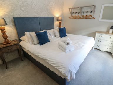 A bedroom with a double bed with blue cushions towels on bed wooden bedside table with lamp and a white dresser with mirror at Apple Tree Cottage in Gweek