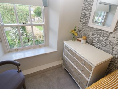 A room corner with a window, a white dresser with decorative items, a mirror, and a chair at Apple Tree Cottage in Gweek