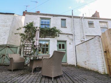 An outdoor patio with wicker chairs a table and an umbrella next to a white stone house with green windows and door at Apple Tree Cottage in Gweek