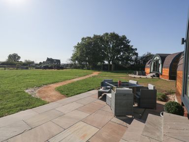 An outdoor patio with table and chairs next to a wooden pod building and a grass lawn with a dirt path at Leadon View Willow in Upleadon near Newent