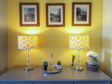 A console table with two decorative lamps a tray with a bottle and plants a vase with flowers and three framed pictures on a yellow wall at Hendre in Blaenau Ffestiniog