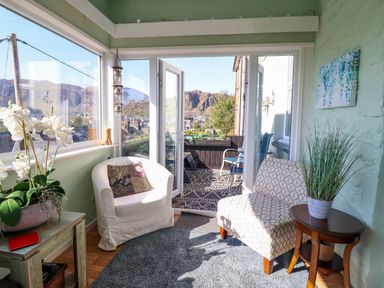 A sunroom with two chairs a side table with a plant and an open door leading to a balcony with a view of mountains at Hendre in Blaenau Ffestiniog