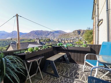 A balcony with chairs a table and a bench with potted plants on the railing overlooking mountains at Hendre in Blaenau Ffestiniog