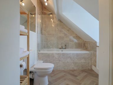A bathroom with a toilet wooden shelving unit with towels a bathtub beneath a slanted ceiling and tiled walls at 47 Church Street in Lyme Regis