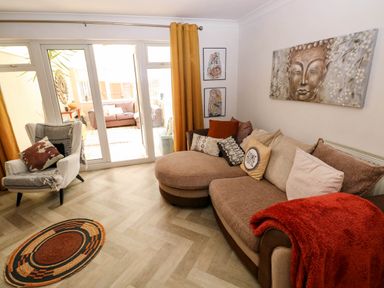 A living room with a brown curved sofa with cushions a white armchair with a blanket and cushions a round rug a glass door with yellow curtains and a sofa visible outside at Alban Road in Llanelli