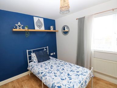 A single bed with blue leaf patterned bedding next to a blue accent wall with a wooden shelf and a window with curtains at Alban Road in Llanelli