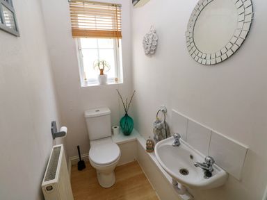 A small bathroom with a toilet, sink, round mirror, window with wooden blinds, and decorative items at Alban Road in Llanelli