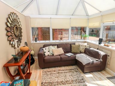 A conservatory with a brown leather sofa and cushions a wooden side table with decorations and a large wall mirror at Alban Road in Llanelli