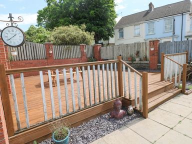 A wooden deck with railing and stairs in a backyard with potted plants and a clock on a wall at Alban Road in Llanelli