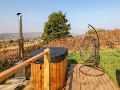A wooden hot tub with a black cover and a metal chimney next to a hanging wicker chair on a wooden deck outside at Holbein in Matlock