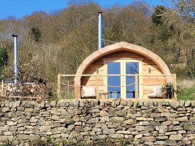 A wooden pod with a curved roof and glass doors with chairs on a small porch behind a stone wall at Holbein in Matlock