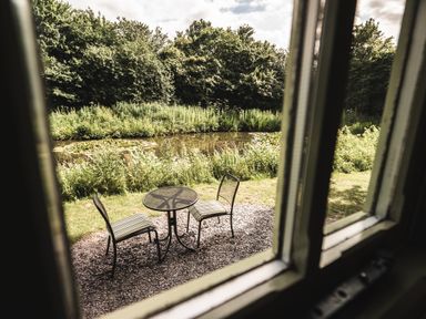 A small outdoor seating area with two striped chairs and a round table by a pond with greenery at Little Otchan Shepherd's Hut in Halsham near Roos