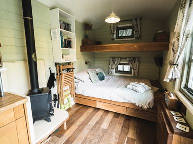 A small bedroom with a wood stove a bed under a wooden loft and shelves at Little Otchan Shepherd's Hut in Halsham near Roos