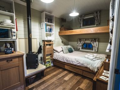 A small bedroom with a bed beneath a lofted wooden shelf two small windows a wood burning stove and built in shelves Little Otchan Shepherd's Hut Halsham near Roos