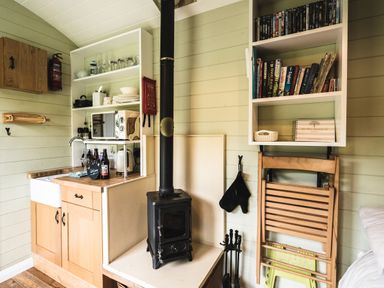 A small kitchen area with wooden cabinets a microwave shelves with dishes a wood stove and a bookshelf with books at Little Otchan Shepherd's Hut Halsham near Roos