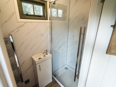 A small bathroom with a corner shower enclosure a white sink unit a window and a chrome towel radiator at Little Otchan Shepherd's Hut Halsham near Roos