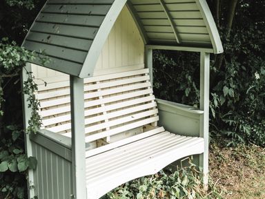 A wooden garden bench with a roof set among plants and bushes at Little Otchan Shepherd's Hut in Halsham near Roos