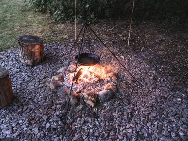 An outdoor fire pit with a tripod and pan over the fire next to wooden log stools