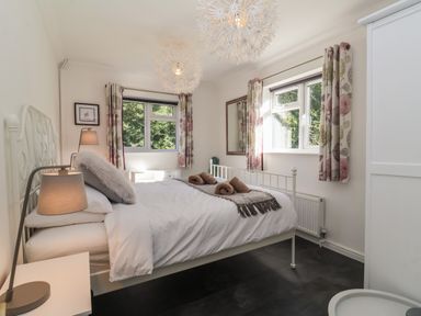 A bedroom with a white metal bed frame bedside tables lamps a large mirror floral curtains and two windows at The Corner House in Ufford near Woodbridge