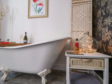 A bathroom with a clawfoot bathtub a wooden tray holding a bottle and glass a side table with a wicker drawer a bottle of bath oil and a glass jar with fairy lights at The Corner House in Ufford near Woodbridge