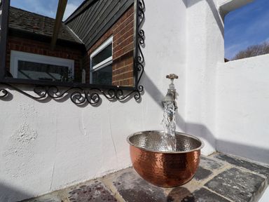 An outdoor water tap with running water into a copper basin under a mirror on a white wall at The Corner House in Ufford near Woodbridge