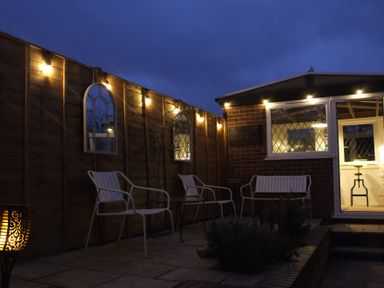 An outdoor patio area with metal chairs and a bench along a wooden fence with decorative mirrors and lights at The Corner House in Ufford near Woodbridge