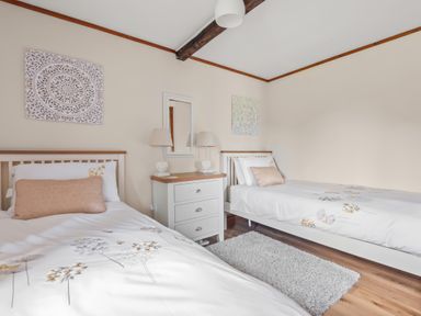 A bedroom with two single beds a white rug a chest of drawers with lamps and wall decorations at The Dairy in St Neot