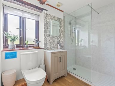 A bathroom with a toilet, a sink with a cabinet, a shower area with glass partition, and a window with plants on the sill at The Dairy in St Neot