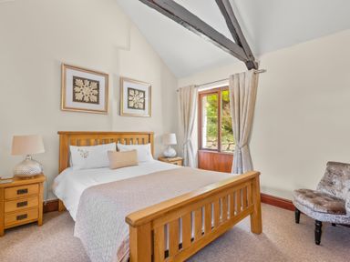 A bedroom with a wooden bed frame side tables lamps wall art a window with curtains and a chair at The Stables in St Neot