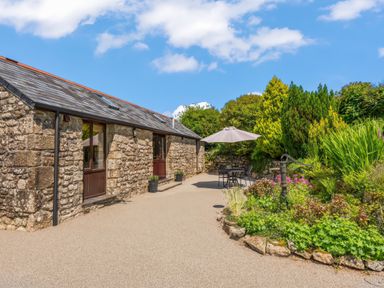 A stone building with wooden doors and windows next to a patio with a table umbrella and chairs and a garden with plants at The Stables in St Neot