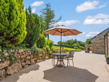 An outdoor patio with a round table, two chairs, and a large umbrella next to a stone wall with bushes at The Stables in St Neot
