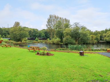 A grassy area with picnic tables next to a pond surrounded by trees at Lakeside Lodge in Bowland Lakes Leisure Village near Garstang