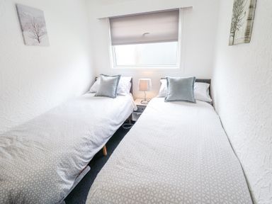 A bedroom with two single beds a window with a blind and a nightstand with a lamp between the beds at 104 The Laurels in Chapel St Leonards
