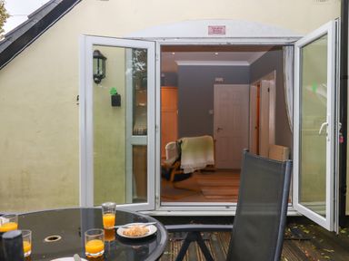 An outdoor patio with a glass table and chairs with orange juice and a croissant in front of open double doors leading to a living room with a chair and a door at 5 Lakeview Rise Highampton near Hatherleigh