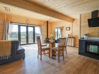 An open dining and kitchen area with wooden walls and ceiling a round dining table with four chairs a grey sofa and large glass door with curtains at Foxglove Higher Wambrook near Chard