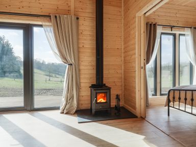 A wood paneled room with a wood burning stove near large glass doors and a bedroom visible through an open doorway at Foxglove Higher Wambrook near Chard