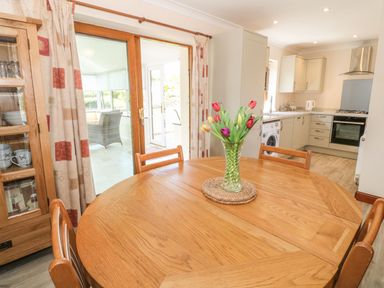A dining area with a wooden table and a vase of flowers next to a kitchen with white cabinets and appliances at Bwthyn Clyd in Llanddaniel Fab