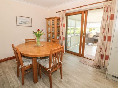 A dining area with a round wooden table and four chairs with cushions a glass cabinet a vase with flowers and glass sliding doors leading to a sunroom at Bwthyn Clyd in Llanddaniel Fab